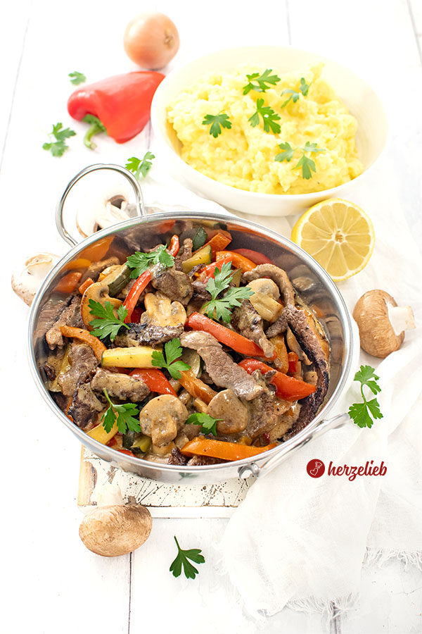 Pfannengericht nach einem Steakpfanne Rezept von herzelieb in einer silberfarbenen Servierpfanne. Im Gericht sieht man Paprika, Zucchni, Teakstreifen und Pilze iner cremigen Senfsoße. Im Hintergrund eine Schüssel mit Kartoffelbrei.